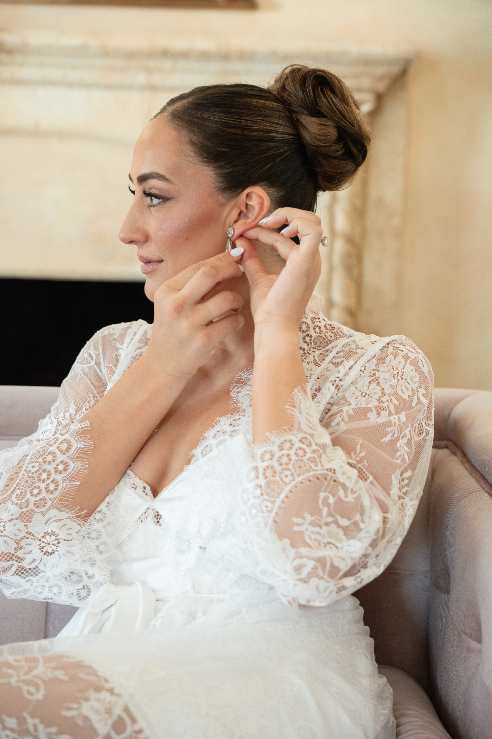 Bride in lace robe putting on earrings, preparing for her wedding.