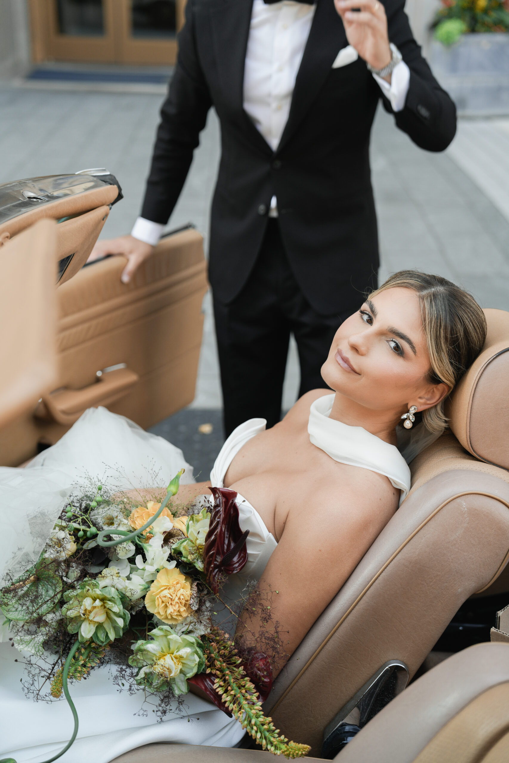 Bride sitting in a car holding a bouquet, dressed in a white wedding gown.
