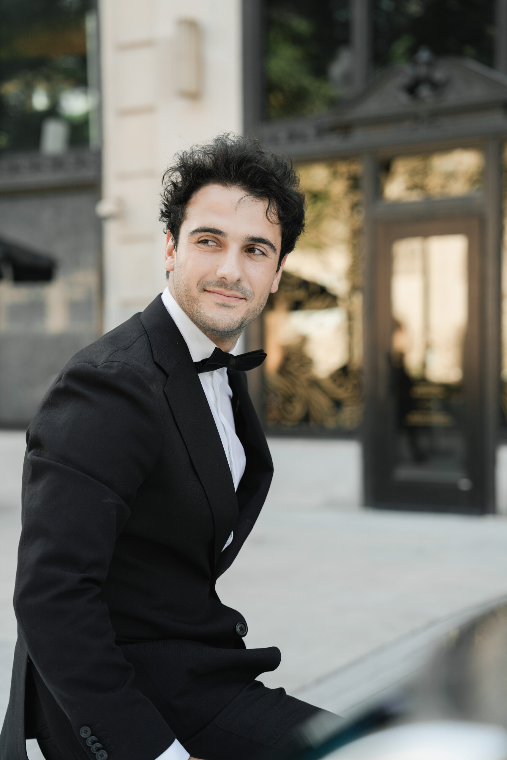 A man in a black tuxedo smiling outdoors.