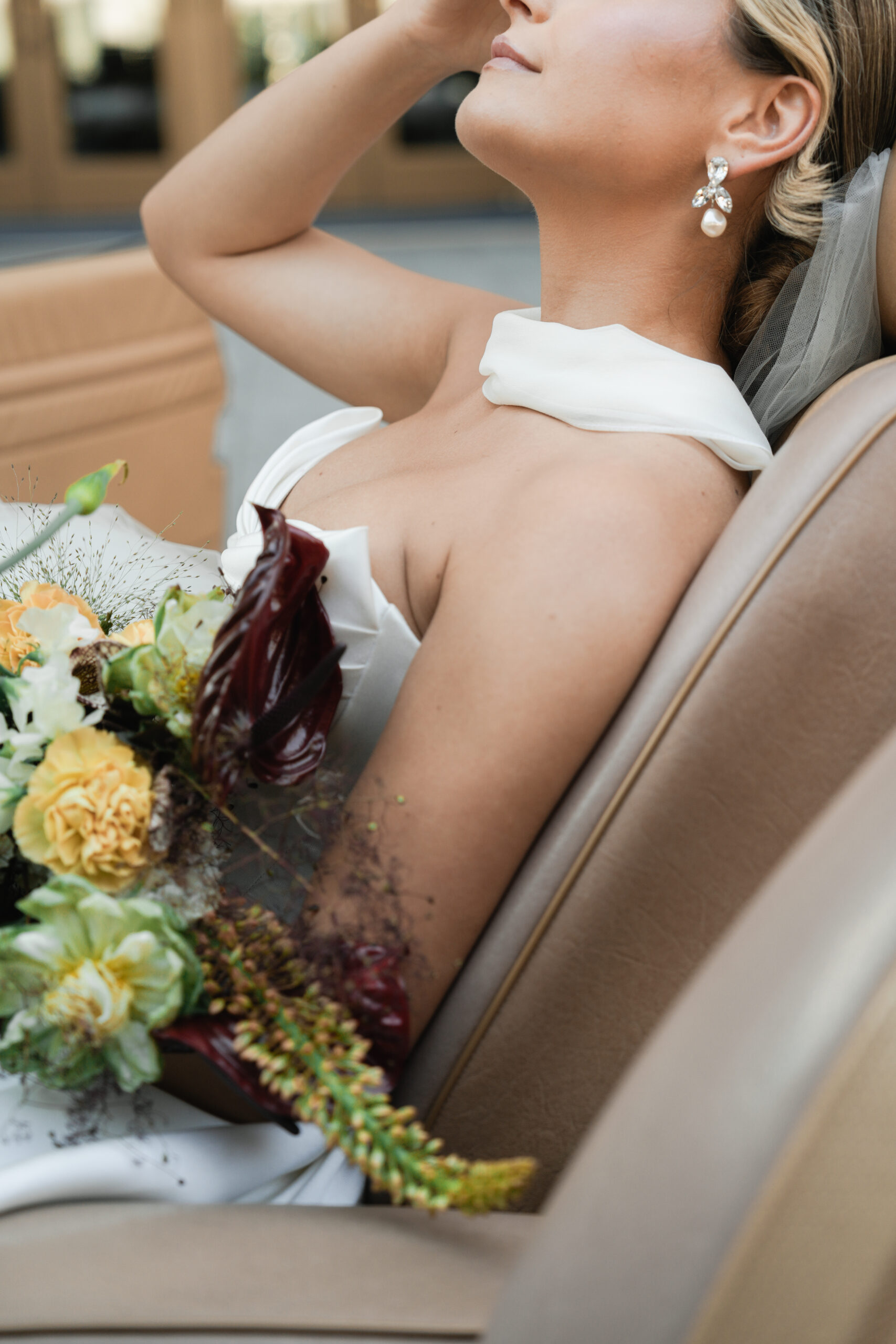 Bride in a white dress holding a bouquet, sitting on a beige chair.