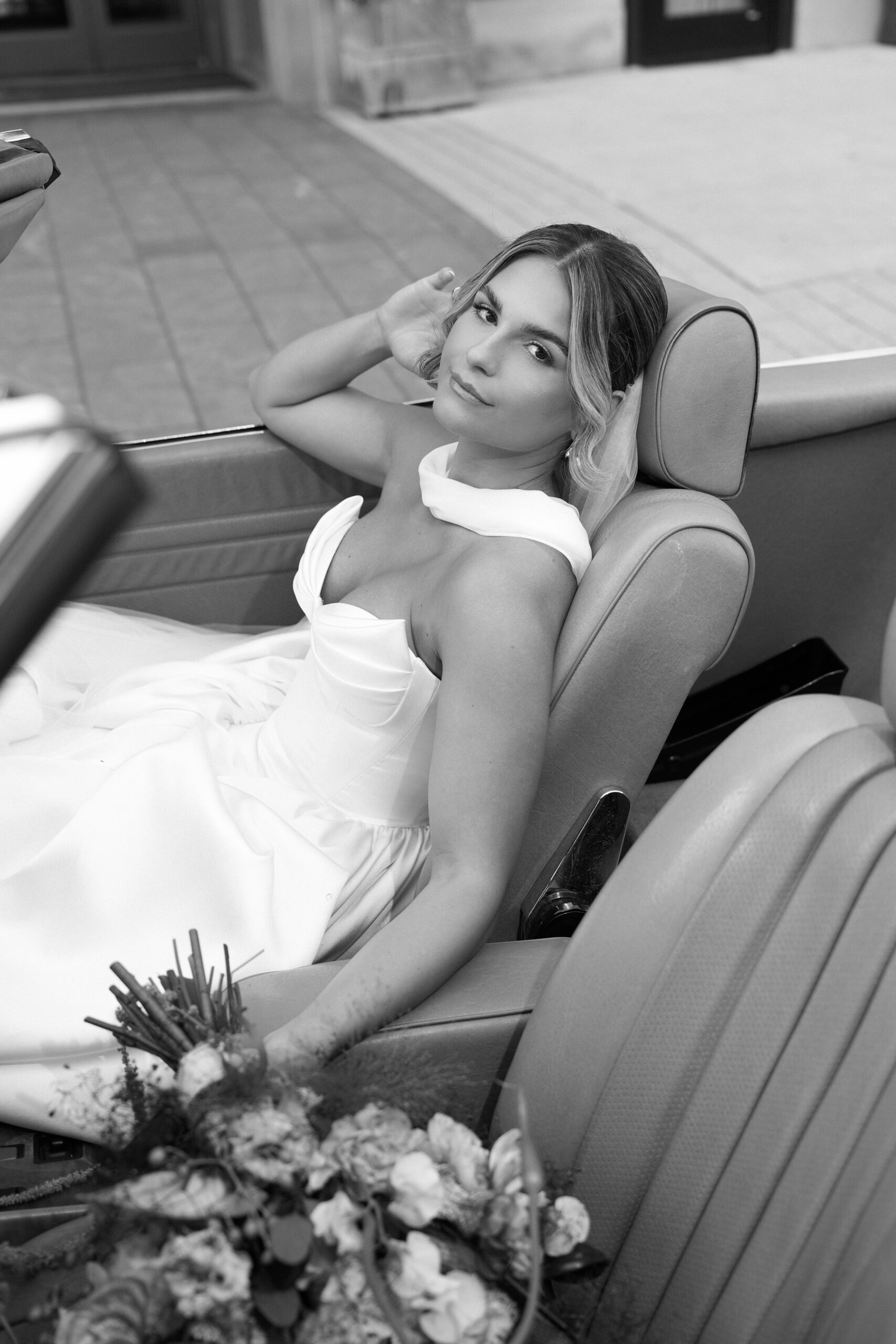Bride in white dress sitting in a car, holding flowers.