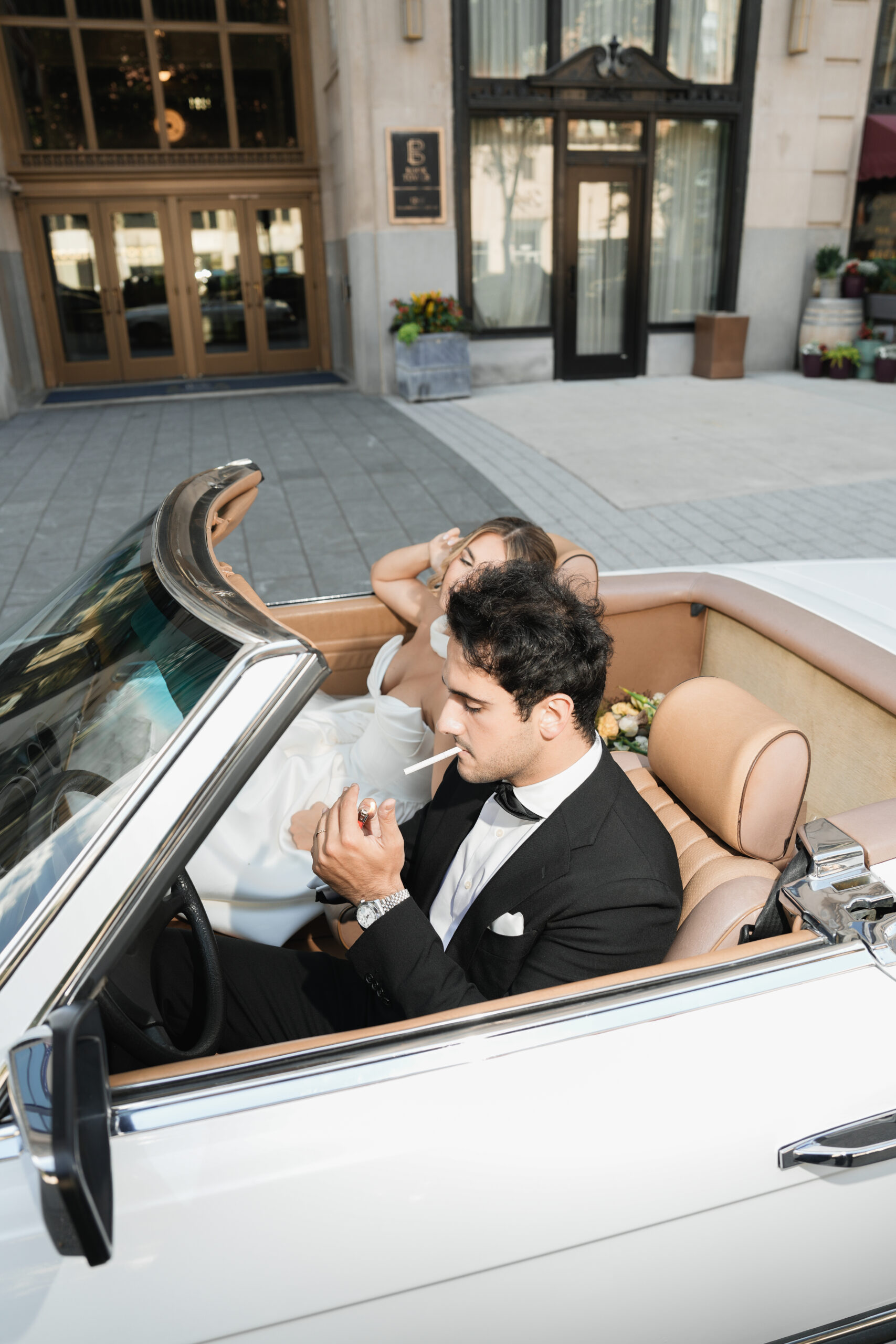 A man in a tuxedo adjusts his watch while sitting in a convertible car.
