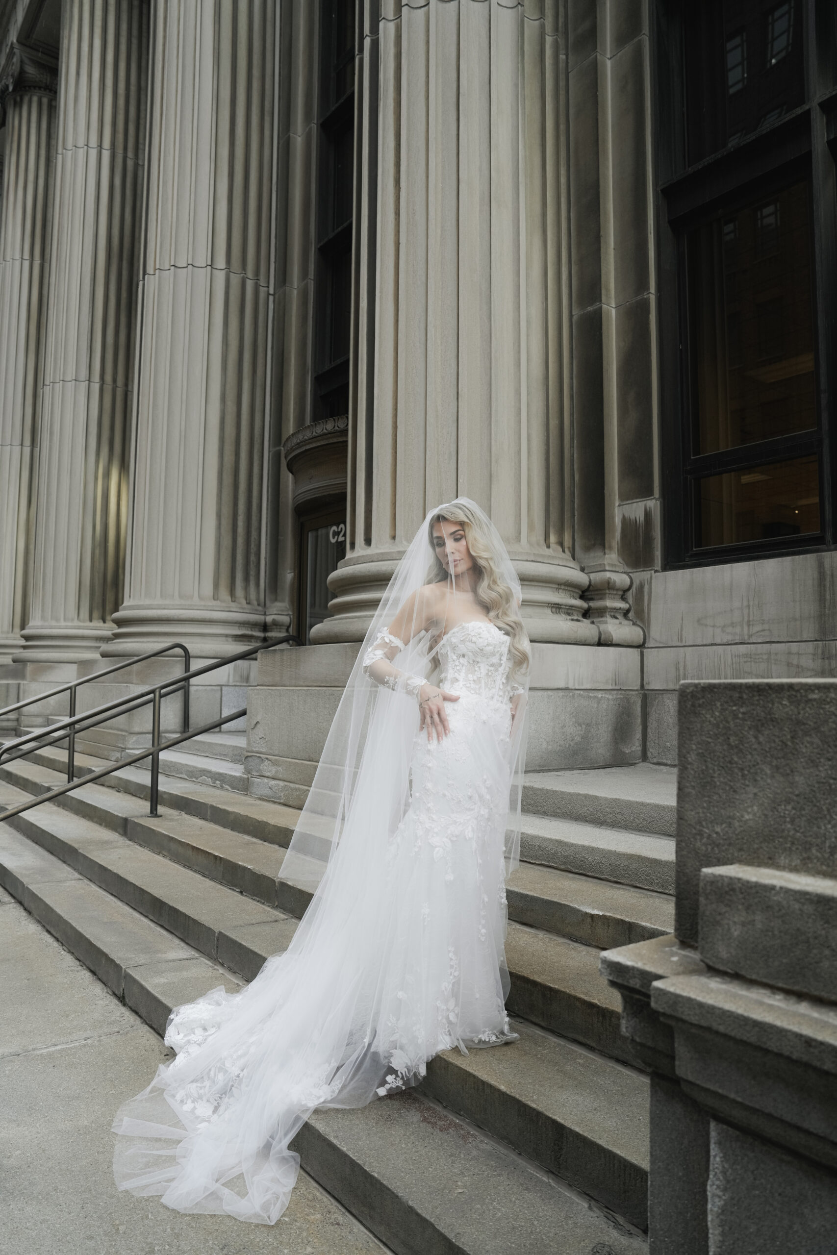 Bride in elegant white gown standing on grand stone steps outside a classical building.