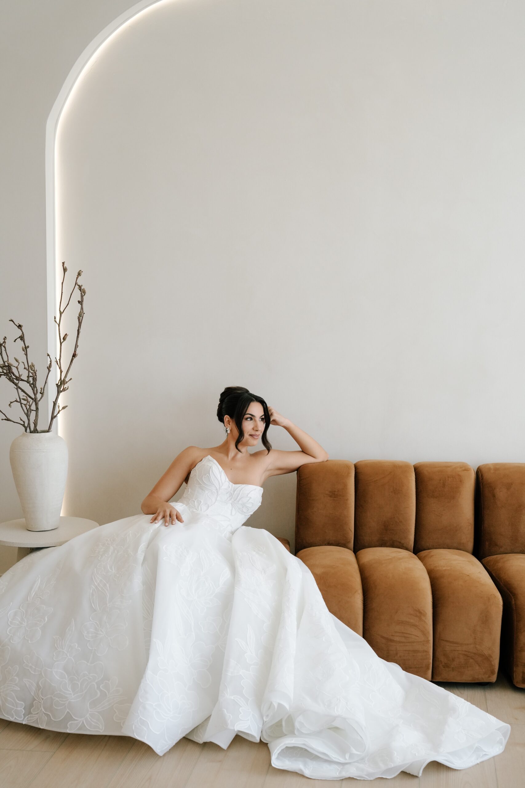 Bride in elegant white gown relaxing on a brown sofa.