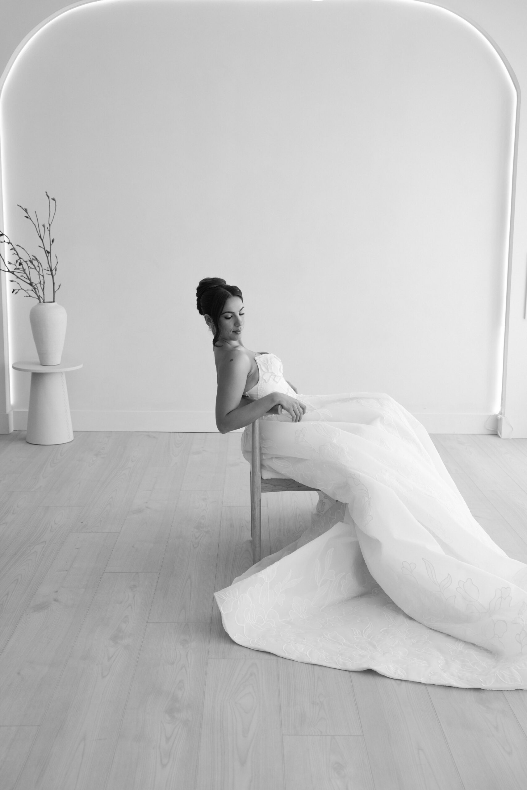 Bride in a flowing wedding gown sits gracefully on a chair in a minimalist room.
