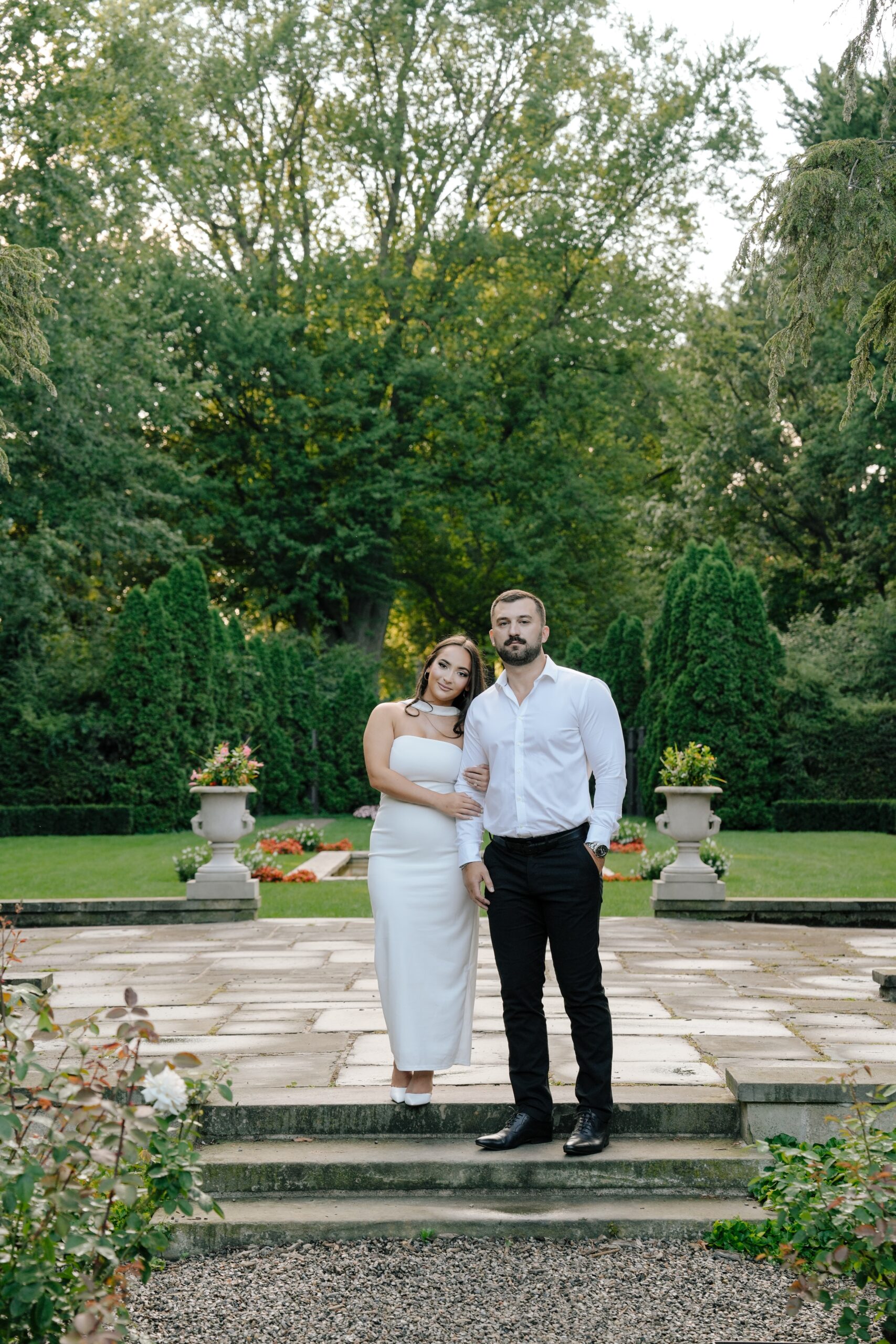 A couple posing outdoors in formal attire surrounded by greenery.